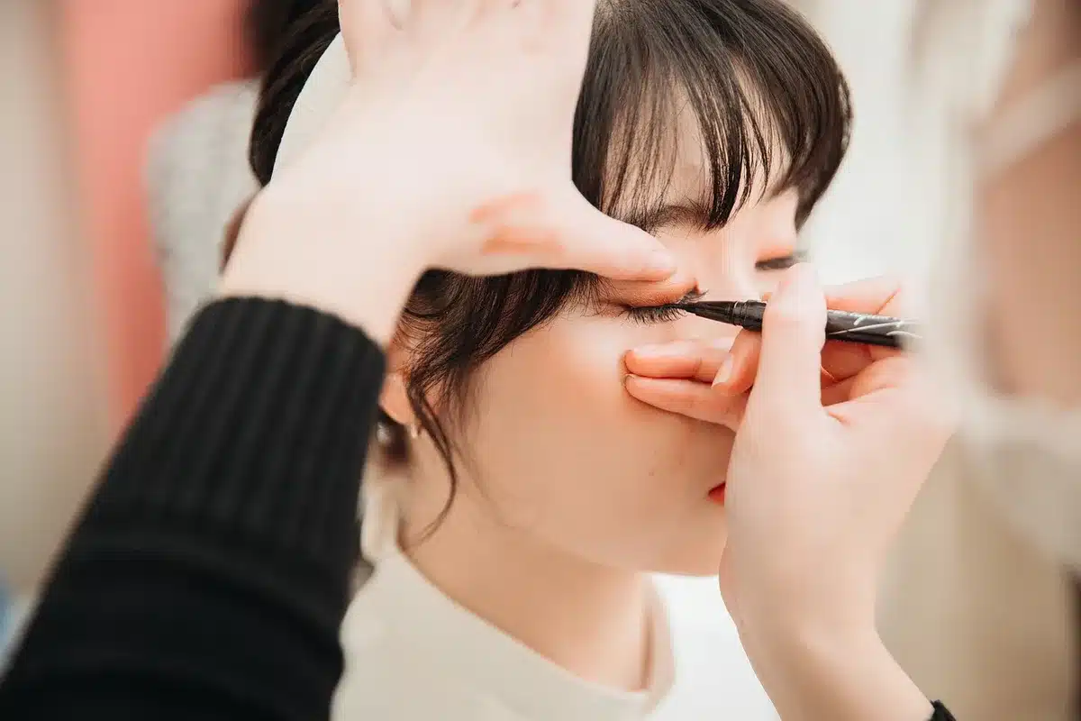Hands holding a compact mirror beside small cosmetic vials on a dressing table in soft daylight